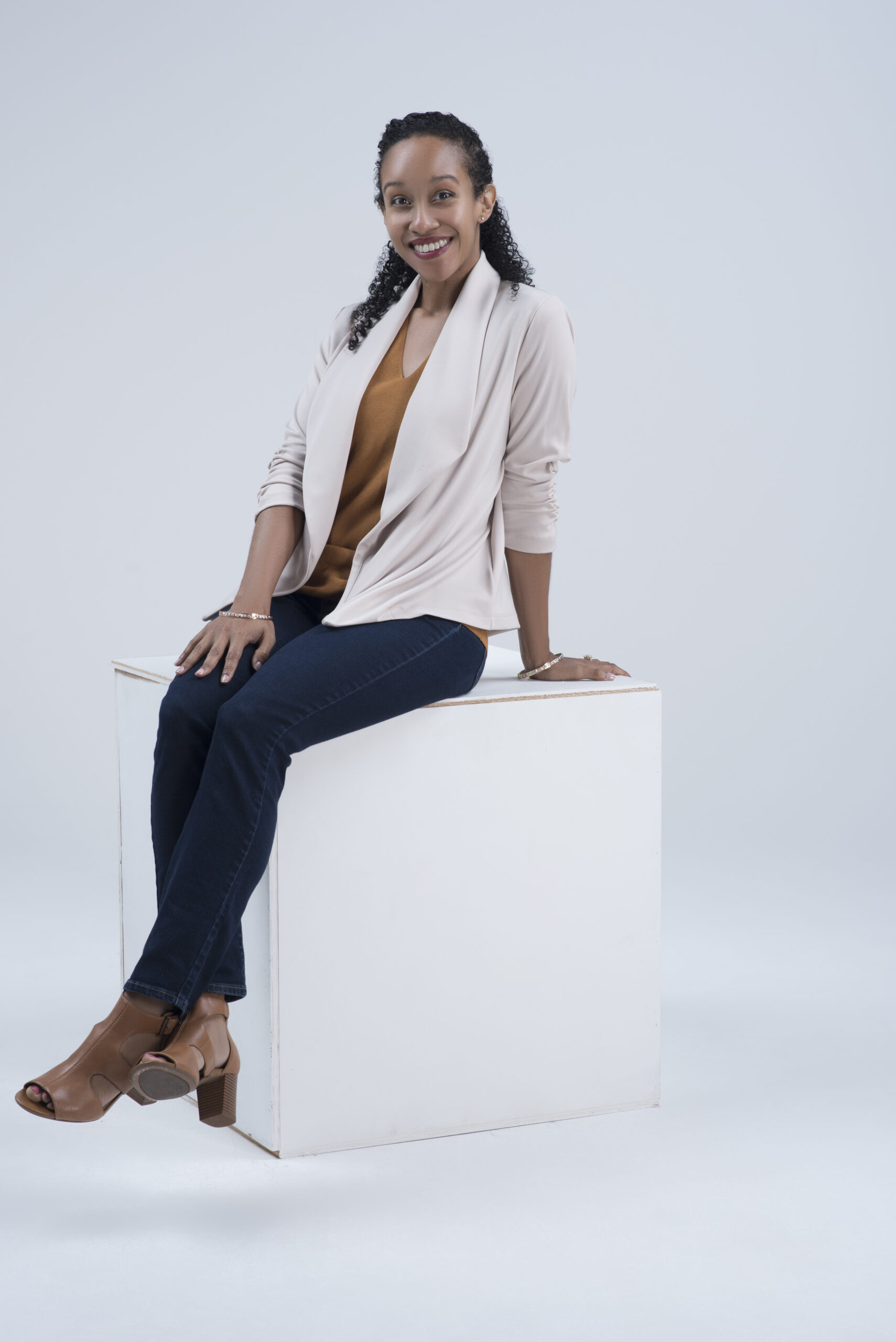 Woman with brown skin and curly hair in a cream jacket, brown blouse, dark pants, and brown heels.
