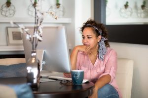 Brown skin woman with brown curly hair, a pink shirt and blue jeans reading from her desktop computer