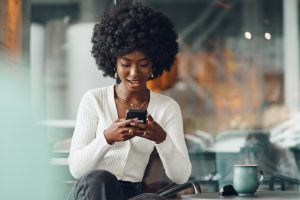 Brown skin woman with curly afro white sweater and grey pants holding a cellphone