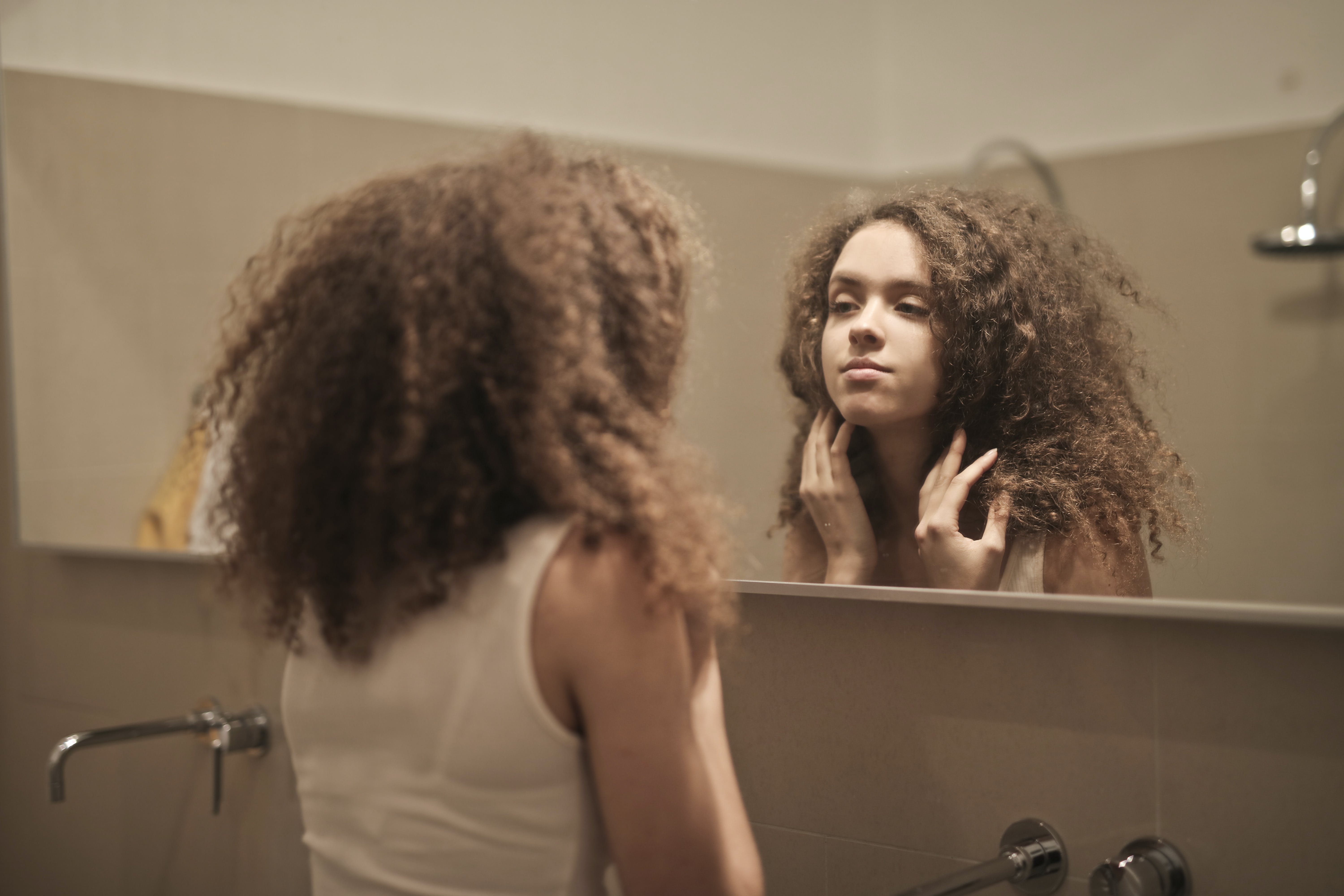 A girl in a pink tank top stands in front of a bathroom mirror and adjusts her curly brown hair. Even comments about physical appearance can affect your child's identity development.