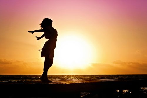 A silhouette of a woman with her arms reaching behind her and looking up at the sky against a sunset background. Womenâ€™s empowerment coaching is meant to help you feel free and confident like this