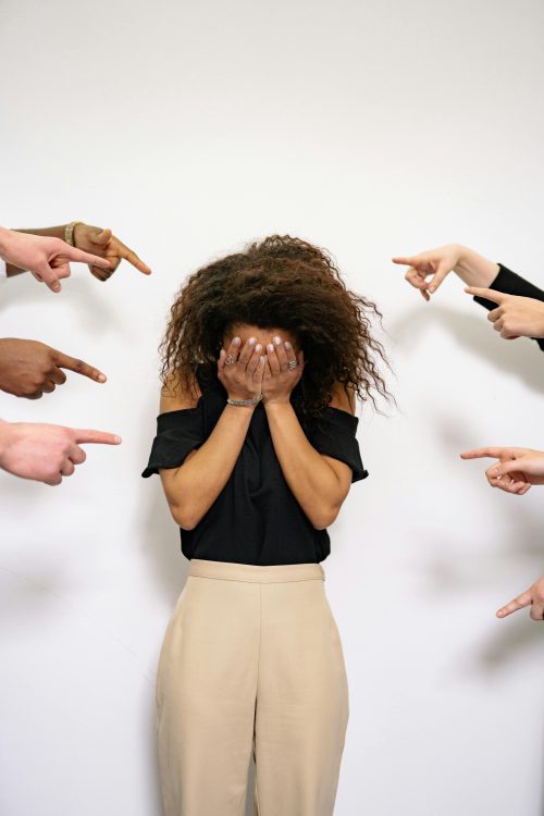 A woman holds her face in her hands, looking distressed as others' hands point at her from both sides. Mixed-race mental health issues often result from feeling like others are pointing at you and your differences constantly.