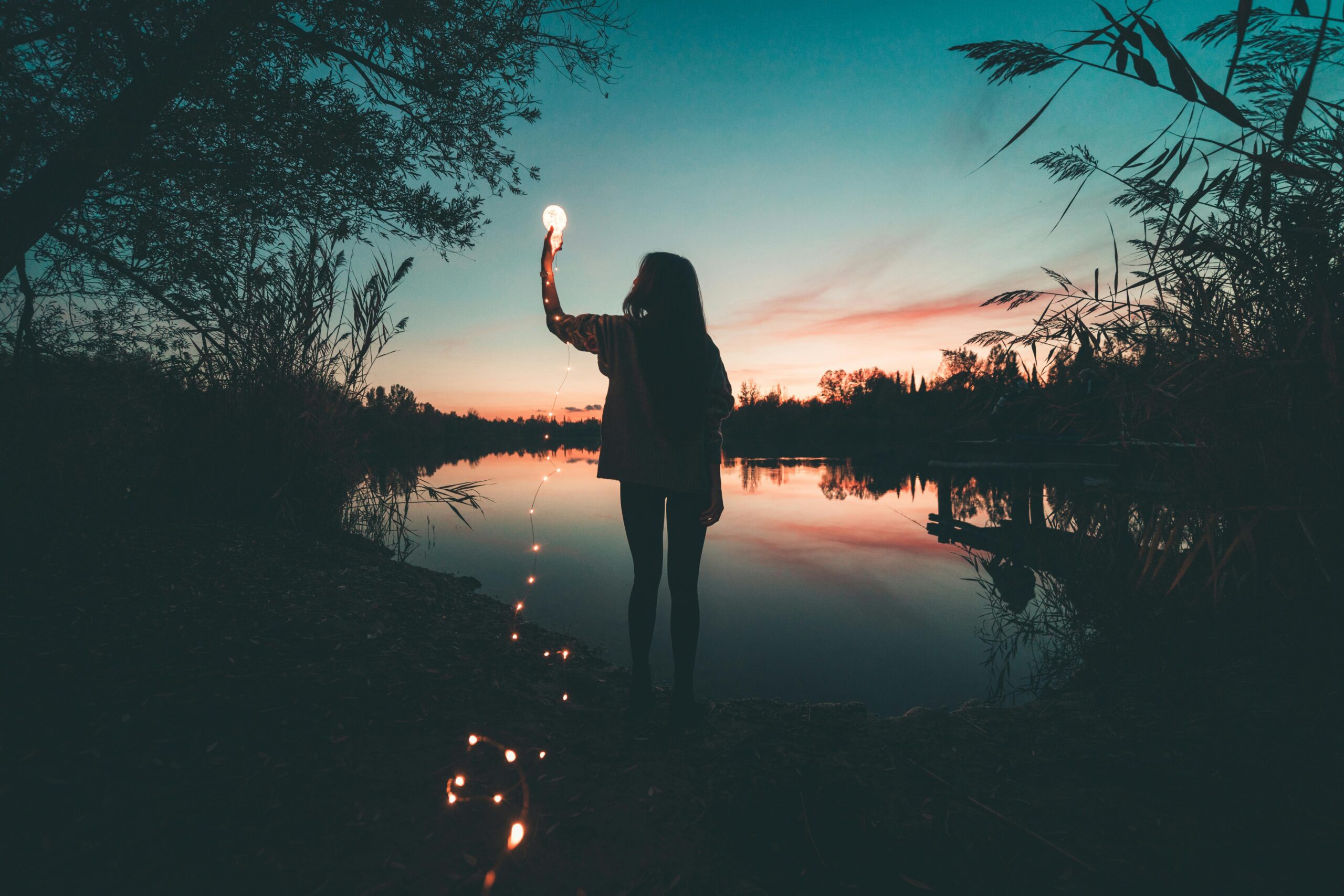 A woman stands in front of a lake and holds a lightbulb up against the night sky, with a string of lights trailing behind her. Pursuing your purpose is shining your inner light for all to see.