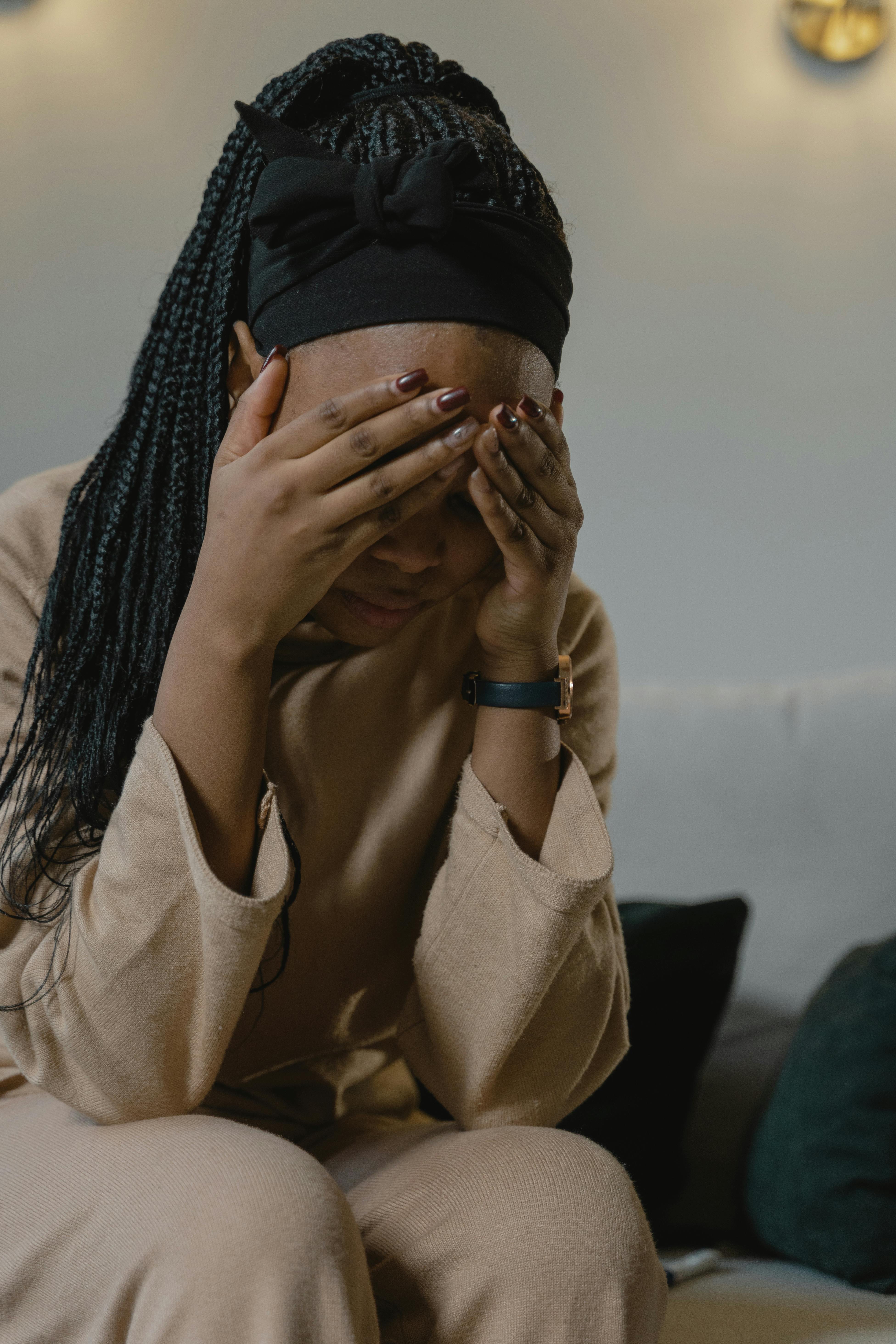 A woman sits on a couch with her head in her heads, feeling overwhelmed by her limiting beliefs