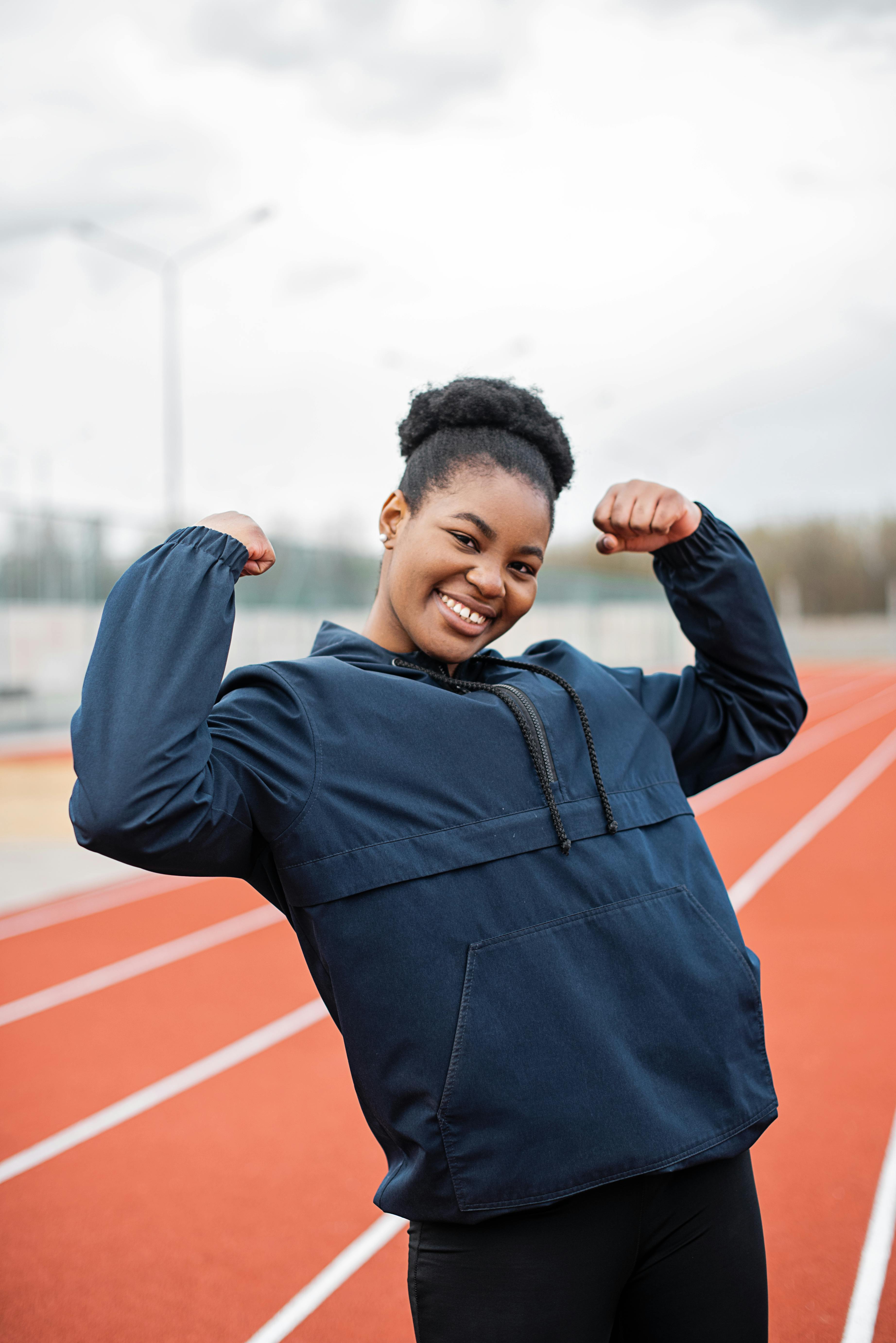 A strong, resilient woman of color stands outside on a running track. She's facing the camera and flexing her arms.