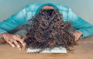 Brown Skin Woman with Blue Shirt and Brown Curly Hair Resting Her Head on a Table