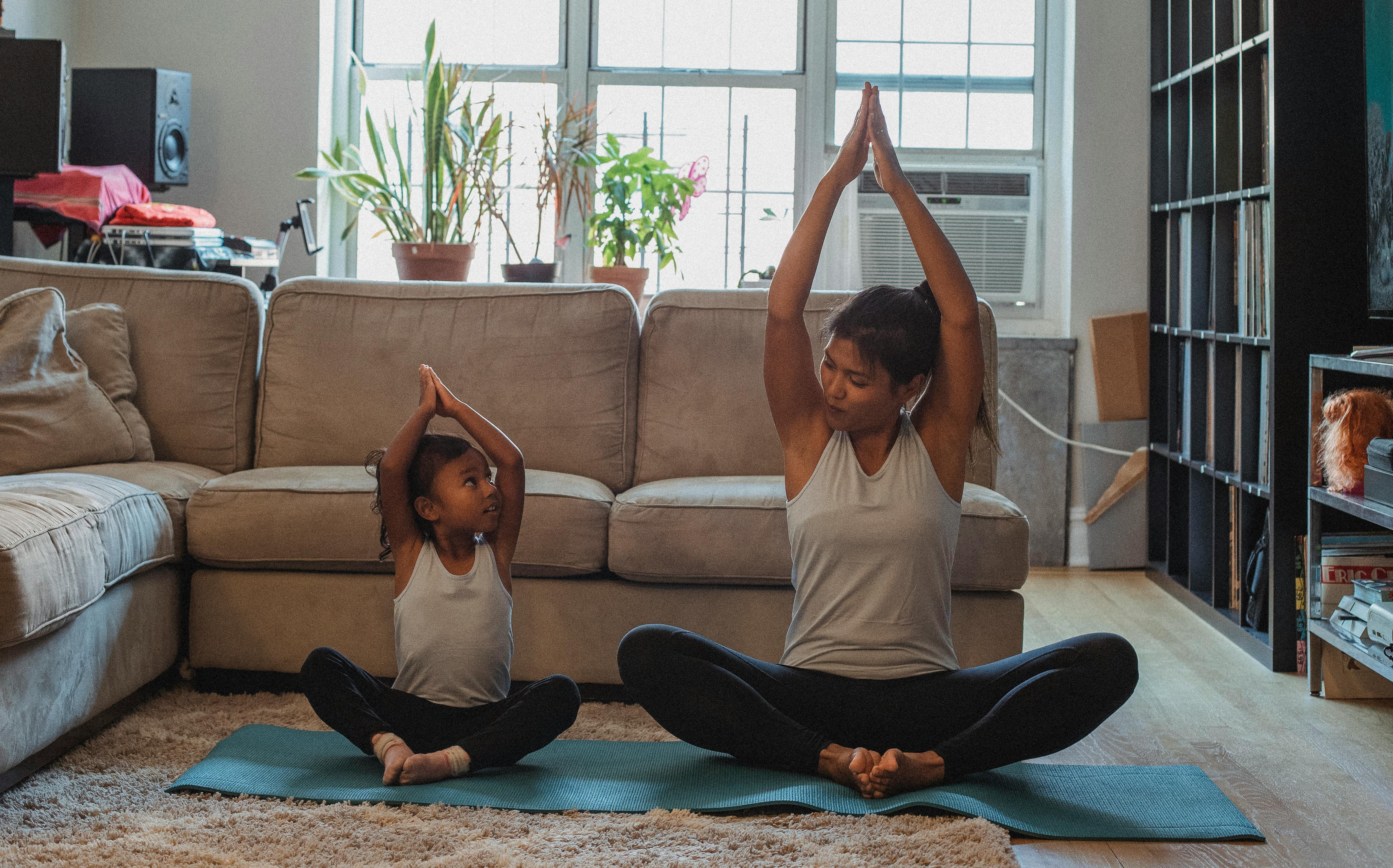 A mom and her daughter sit next to each other in a lotus position on a yoga mat in their home. They're looking at each other with their arms raised above their heads.