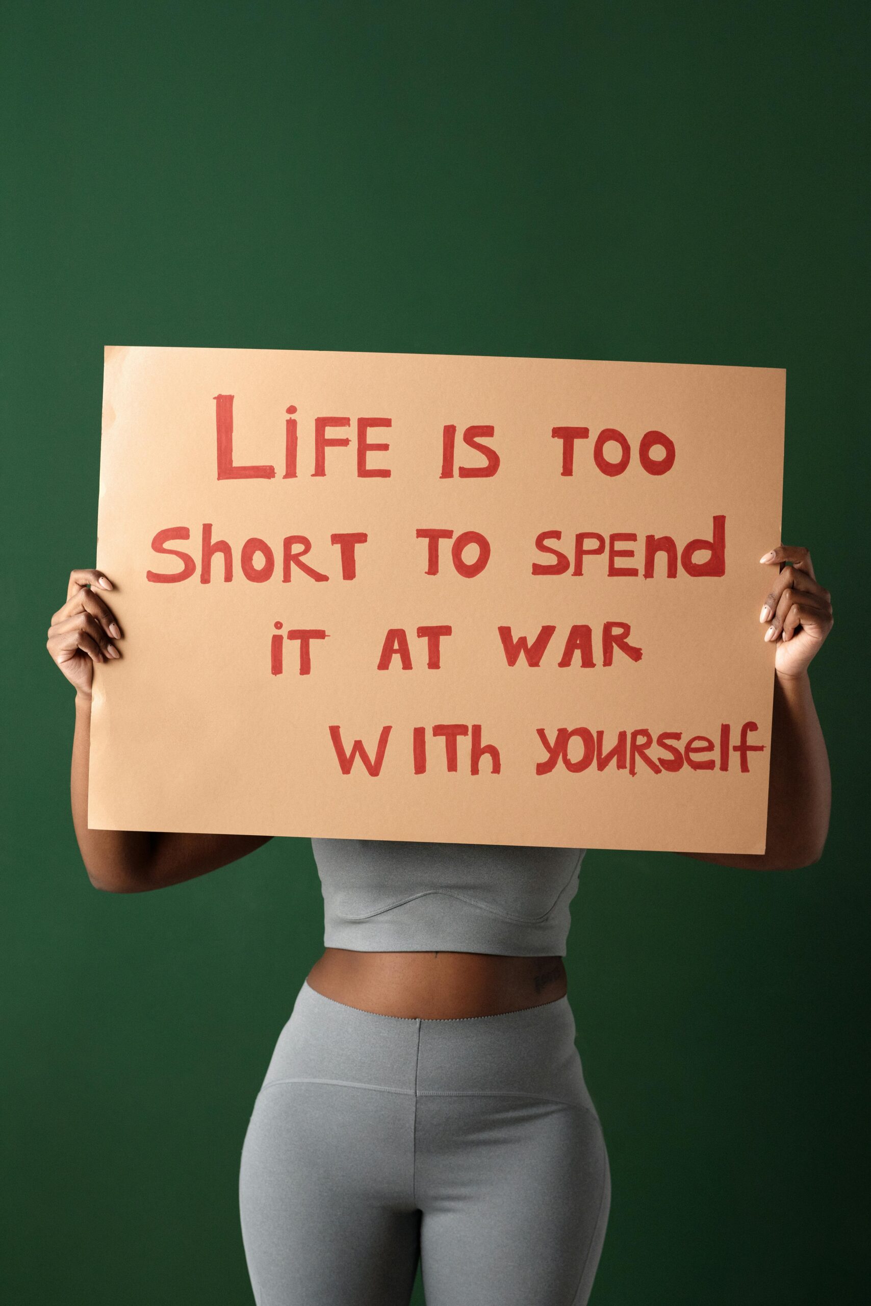 A lady in a two-piece gray outfit holds a sign in front of her face that reads "Life is too short to spend it at war with yourself". It's essential that you learn how to like yourself if you want to be happy."