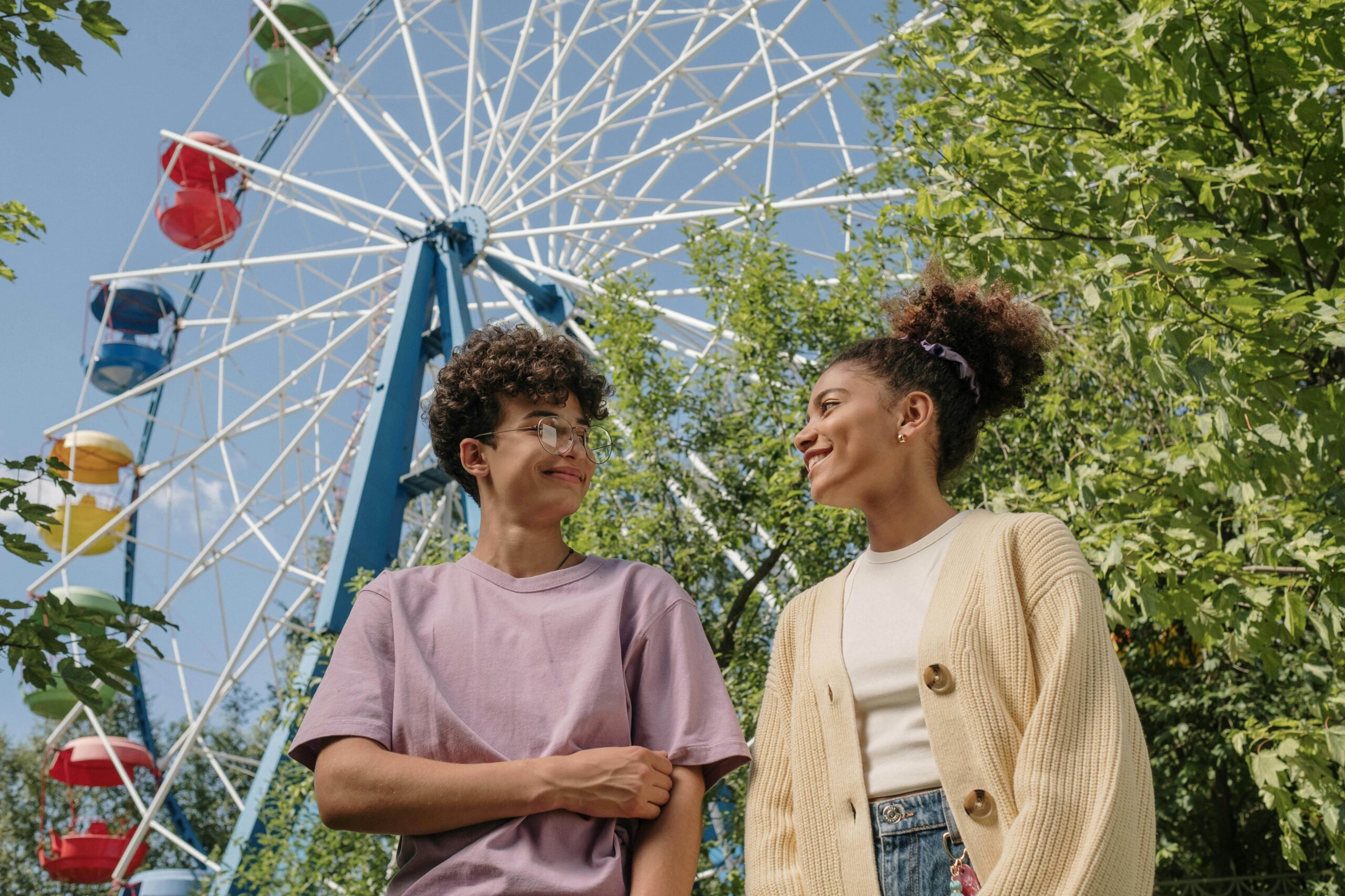 Two mixed teens stand in front of a Ferris wheel smiling at each other.