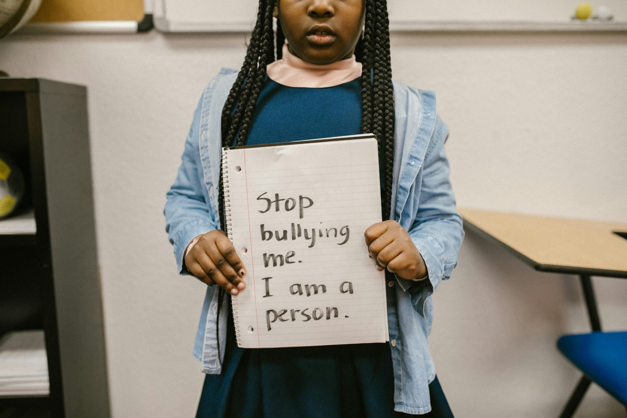 A little girl stands in a classroom holding a sign that says 'Stop bullying me. I am a person.' in a stand against racial bullying.