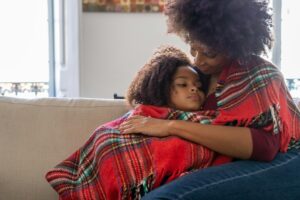 Black mom embracing her mixed daughter beneath a red green and while blanket on a tan couch
