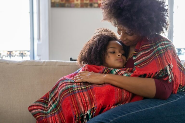 Black mom embracing her mixed daughter beneath a red green and while blanket on a tan couch