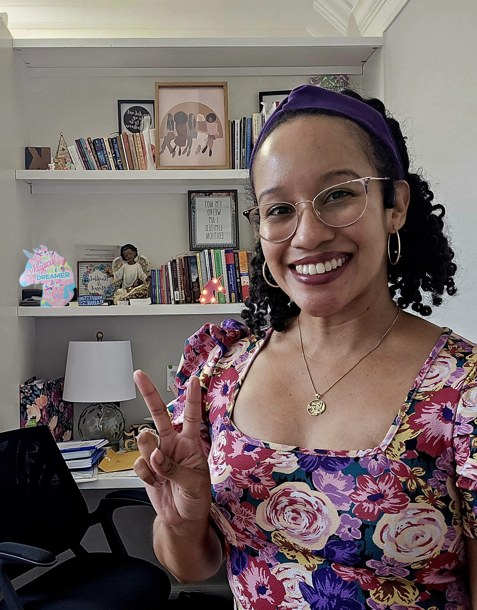 Dr. KayLa wearing a colorful floral blouse, curly dair shoulder-length hair with a purple headband> She is standing in front of a bookcase with various statues, books, and sayings in picture frames