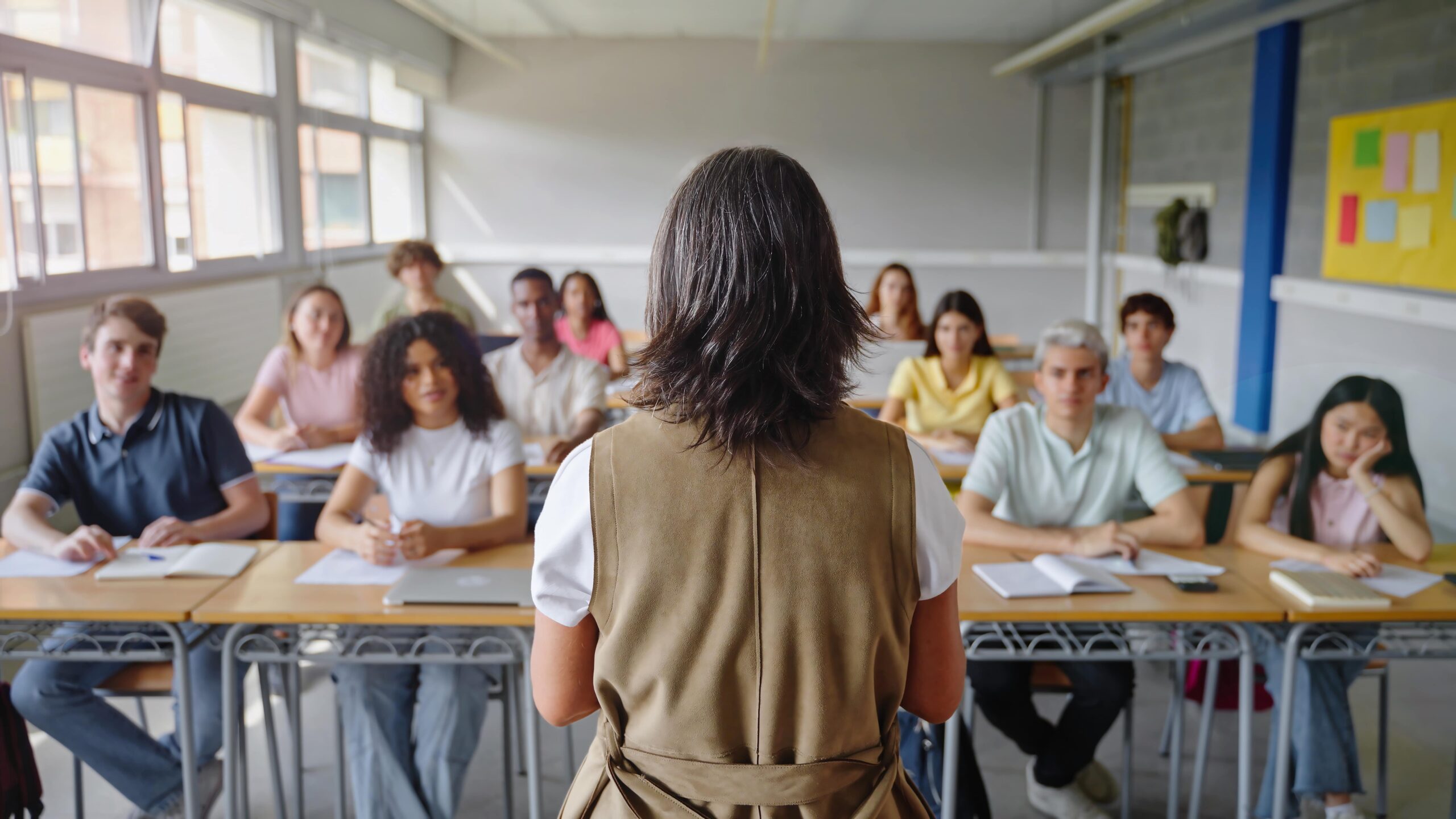 Teacher with short brown and gray hair, brown sleeves vest and white shirt viewing and talking to diverse group teens at classroom desks.