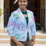 Dr. KayLa N. Allen-Young, a woman with warm medium-brown skin, dark brown eyes, and dark naturally curly hair pulled neatly back with a soft pink ribbon, smiles directly at the camera in an outdoor professional portrait. Her hair is smoothed away from her face and gathered behind her head, and she wears large gold hoop earrings. She is dressed in a fitted white blazer covered with pink floral blossoms and green leaves over a deep emerald-green top, paired with taupe-brown trousers. She stands with a relaxed, confident posture, one hand resting along her thigh and the other near her hip. Behind her, softly blurred dark wooden doors and light stone columns frame the portrait while bright natural daylight illuminates her face and clothing.