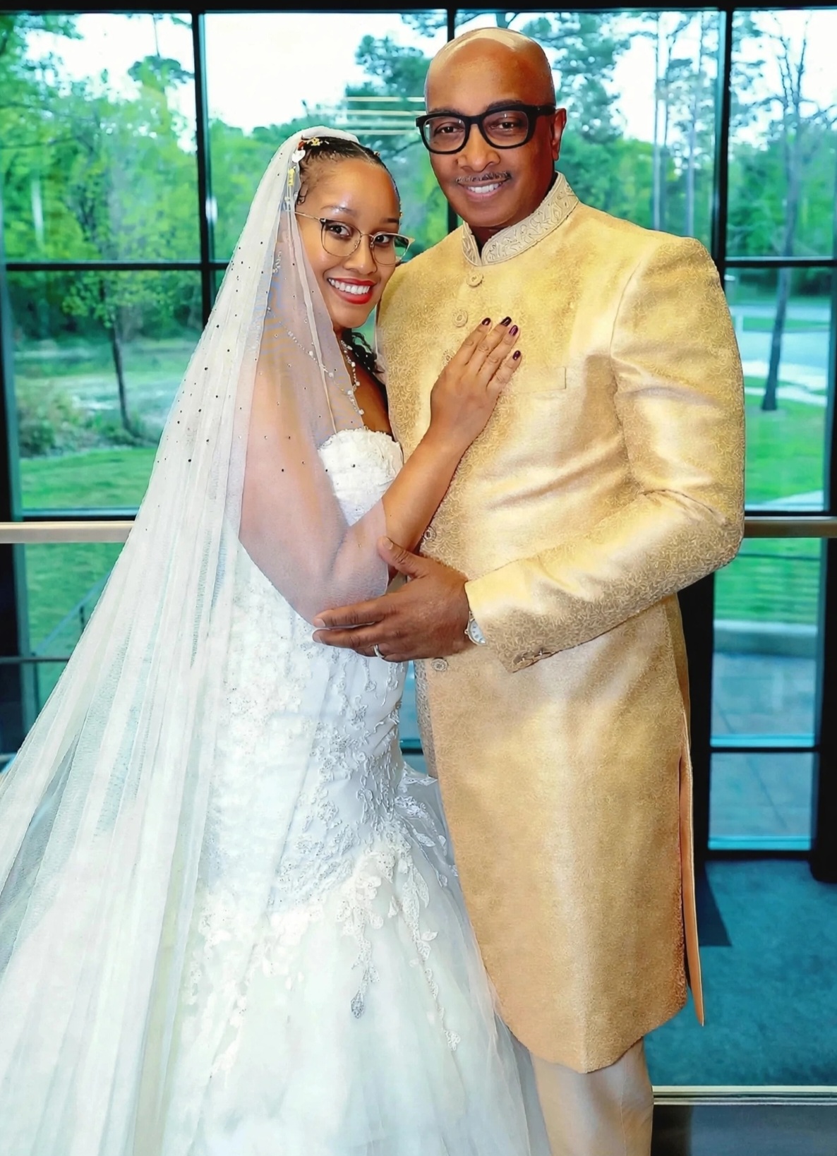 A bride and groom with medium-brown skin smile while standing closely together indoors before tall black-framed windows overlooking lush green trees. The bride wears glasses, red lipstick, a white lace wedding gown, and a sheer veil decorated with red, green, and gold rhinestones. Her hand rests on the groom’s chest, showing burgundy nails and her wedding ring. The groom wears black glasses and an embroidered gold formal suit, with one arm wrapped around her waist.